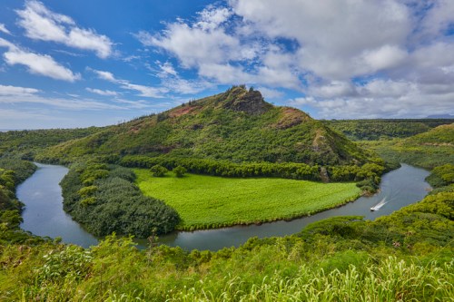 Lush green hills surround a winding river under a blue sky with scattered clouds. A serene landscape scene.