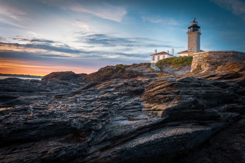 A lighthouse stands on rocky cliffs at sunset, with a colorful sky and calm waters in the background.
