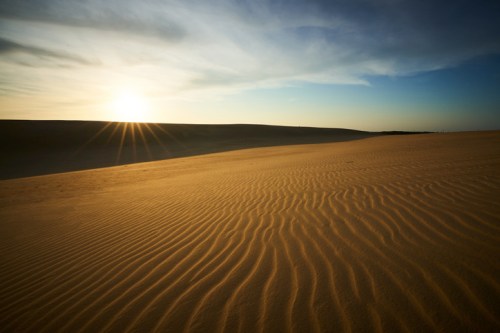 A serene desert landscape at sunset, with rippled sand dunes and a colorful sky.
