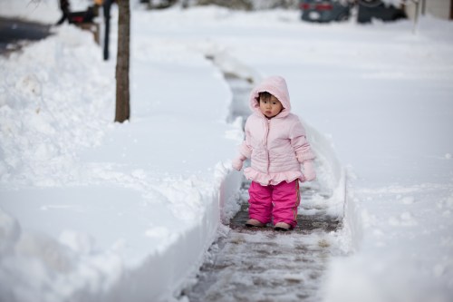A young child in a pink coat and pants walks along a snow-covered path, surrounded by deep snow.