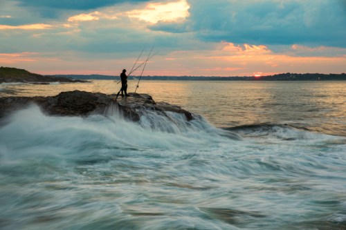 A lone fisherman stands on a rocky shore, casting lines into turbulent waves under a colorful sunset sky.