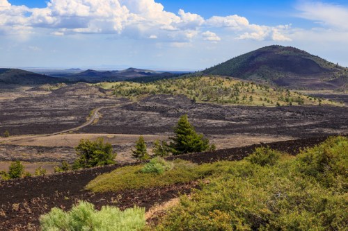 A scenic view of a volcanic landscape with rolling hills, sparse vegetation, and a blue sky dotted with clouds.