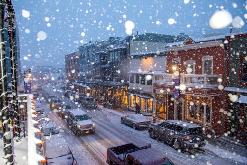 A snowy street scene with cars and shops, illuminated by festive lights, creating a cozy winter atmosphere.