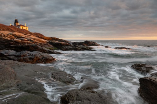 A lighthouse on rocky shores at sunset, with waves crashing against the rocks under a cloudy sky.