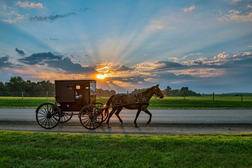 A horse-drawn carriage travels along a road at sunset, with rays of light breaking through the clouds.