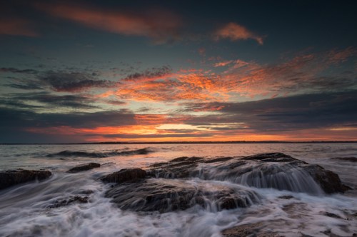 A serene seascape at sunset, featuring rocky shores and vibrant clouds reflecting orange and purple hues in the sky.