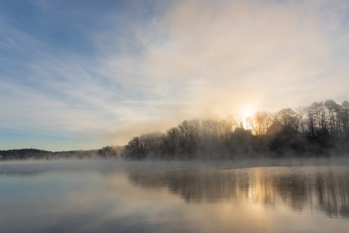 A serene lake at sunrise, shrouded in mist, with trees silhouetted against the soft morning light.