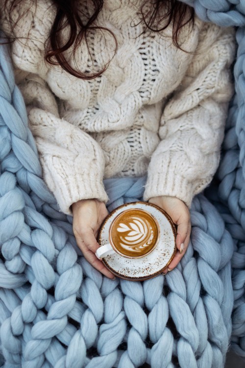 A person in a cozy sweater holds a latte with latte art, sitting under a soft blue blanket.
