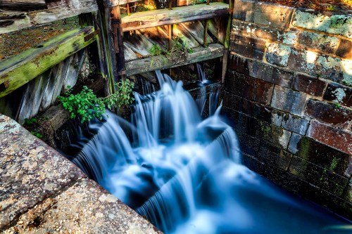 A serene waterfall cascades over stone steps, surrounded by greenery and weathered wooden structures.