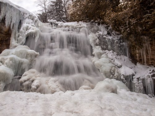 A frozen waterfall cascading over icy rocks, surrounded by snow and frosty trees.