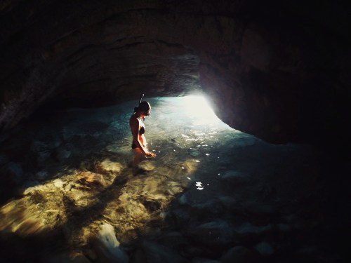 A person stands in shallow water inside a cave, illuminated by light streaming in from an opening above.