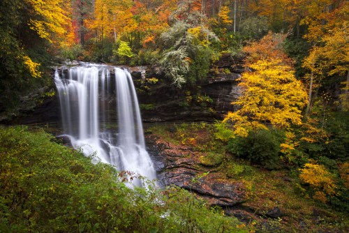 A serene waterfall cascades down rocky cliffs, surrounded by vibrant autumn foliage in shades of orange and yellow.