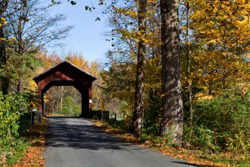 A red covered bridge spans a road surrounded by trees with vibrant autumn foliage.