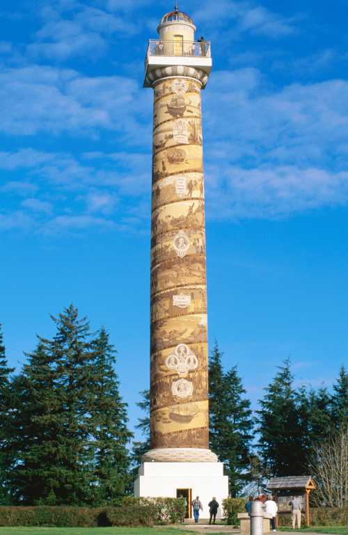 A tall, decorated lighthouse tower surrounded by trees under a blue sky, with people near its base.