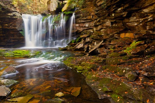 A serene waterfall cascades over rocky cliffs into a calm pool, surrounded by lush greenery and autumn leaves.