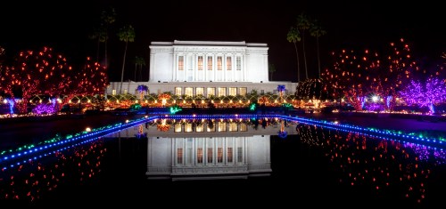 A beautifully lit building at night, reflecting in a pond surrounded by colorful holiday lights and trees.