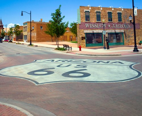 A view of a street in Winslow, Arizona, featuring a large Route 66 sign on the pavement and historic buildings nearby.