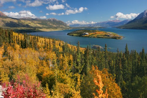 A scenic view of a lake surrounded by vibrant autumn foliage and mountains under a blue sky with fluffy clouds.