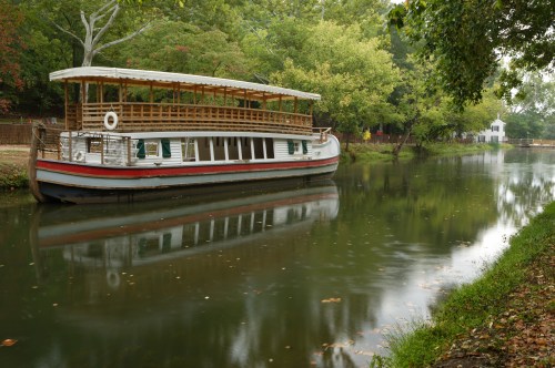 A boat docked on a calm river, surrounded by lush greenery and trees, with a house visible in the background.