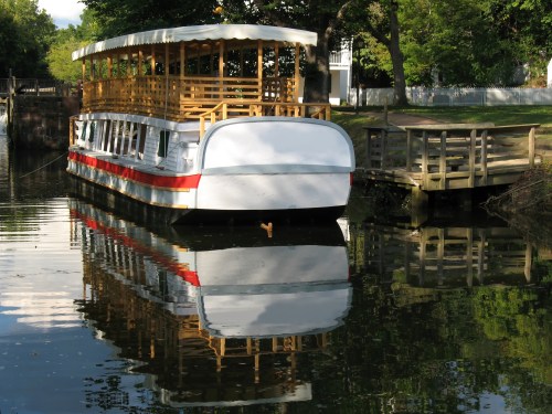 A white and red boat with a wooden deck is moored by a tree-lined canal, reflecting in the calm water.