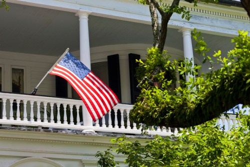 An American flag waves from a balcony, surrounded by green leaves and a white-columned porch.