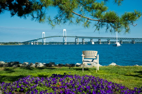A serene view of a bridge over water, with a chair in the foreground and purple flowers lining the shore.
