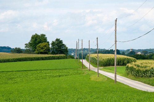 A rural landscape featuring green fields, a winding road, and utility poles under a partly cloudy sky.
