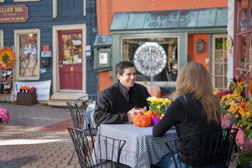 A man and woman sit at a table with flowers, chatting in a colorful outdoor café surrounded by shops.