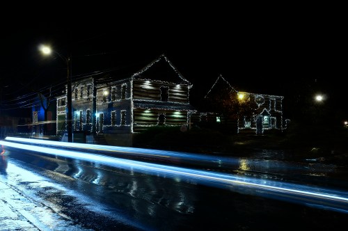 Two houses decorated with Christmas lights at night, with streaks of light from passing cars on a wet road.