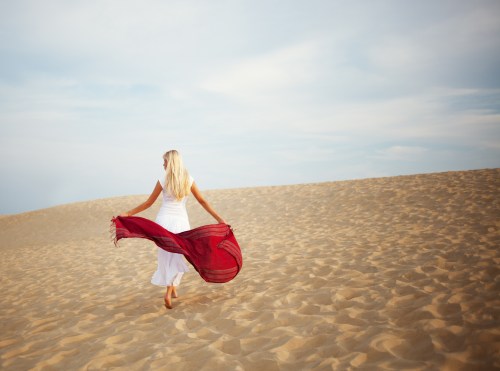 A woman in a white dress walks barefoot on sandy dunes, holding a flowing red scarf against a cloudy sky.
