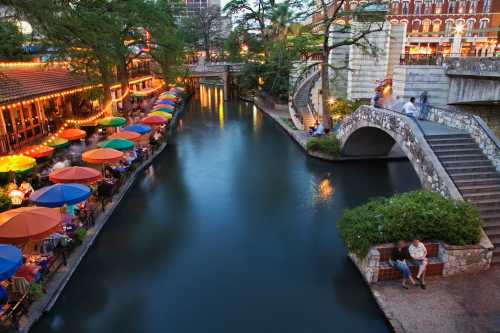 A scenic view of a riverwalk with colorful umbrellas, restaurants, and a stone bridge, illuminated by evening lights.