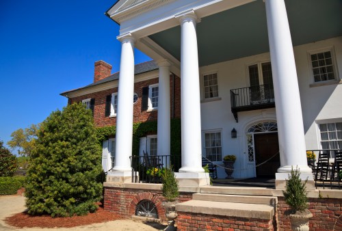A historic building with white columns, brick walls, and a landscaped entrance under a clear blue sky.