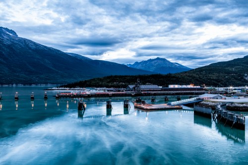 A serene harbor scene with mountains in the background, reflecting clouds and lights on calm water.