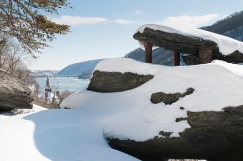 Snow-covered rocks overlook a river and distant hills, with a church steeple visible in the background.