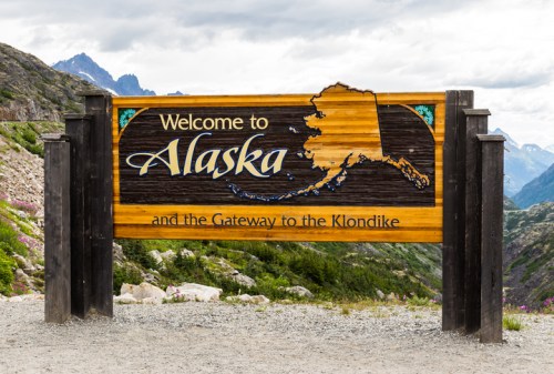 Wooden welcome sign for Alaska, featuring the state outline and text about the Klondike, set against a mountainous backdrop.