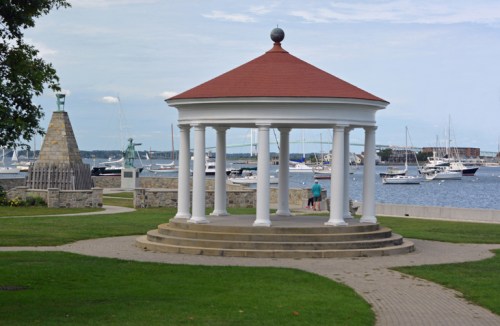 A gazebo with a red roof stands by the water, surrounded by boats and a stone monument in a scenic park.