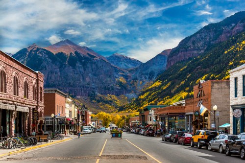 A scenic view of a mountain town with colorful autumn trees, shops lining the street, and majestic mountains in the background.