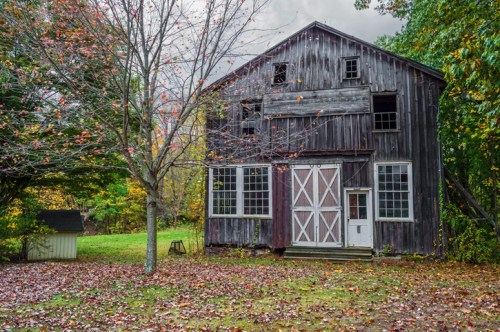 A weathered barn with a white door, surrounded by autumn foliage and fallen leaves on the ground.