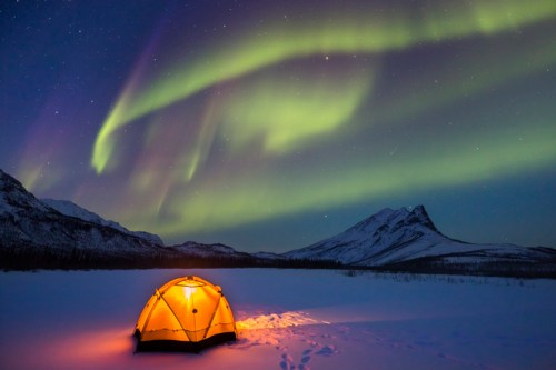 A glowing tent in a snowy landscape under vibrant northern lights and a starry sky. Mountains in the background.