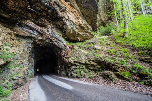 A winding road leads through a rocky tunnel, surrounded by lush greenery and towering cliffs.