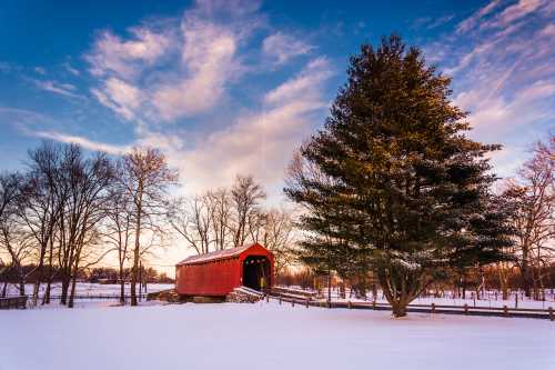 A red covered bridge stands in a snowy landscape, surrounded by bare trees and a colorful sky at sunset.