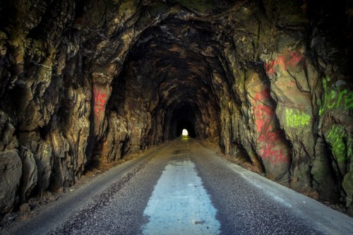 A dark tunnel with rocky walls and colorful graffiti, leading to a bright light at the far end.