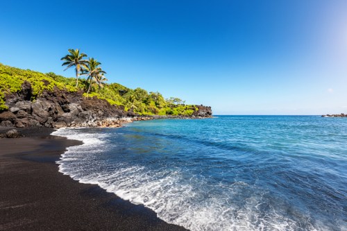 A serene beach with black sand, gentle waves, and lush greenery under a clear blue sky.