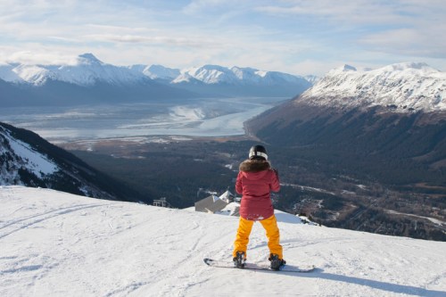 A person in ski gear stands on a snowy mountain, overlooking a valley and distant mountains under a clear sky.