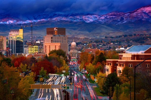 A city skyline at dusk with mountains in the background, colorful autumn trees, and light trails from moving traffic.
