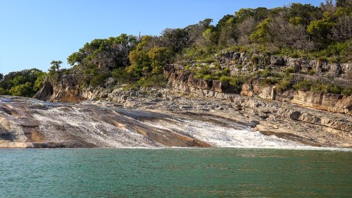A serene landscape featuring a rocky shoreline with cascading water and lush greenery under a clear blue sky.
