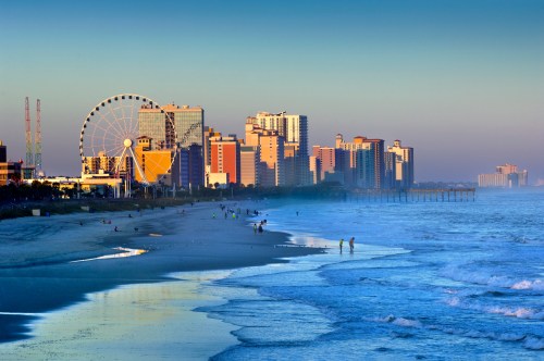 A serene beach scene at sunset, featuring a ferris wheel and city skyline along the shore with gentle waves.