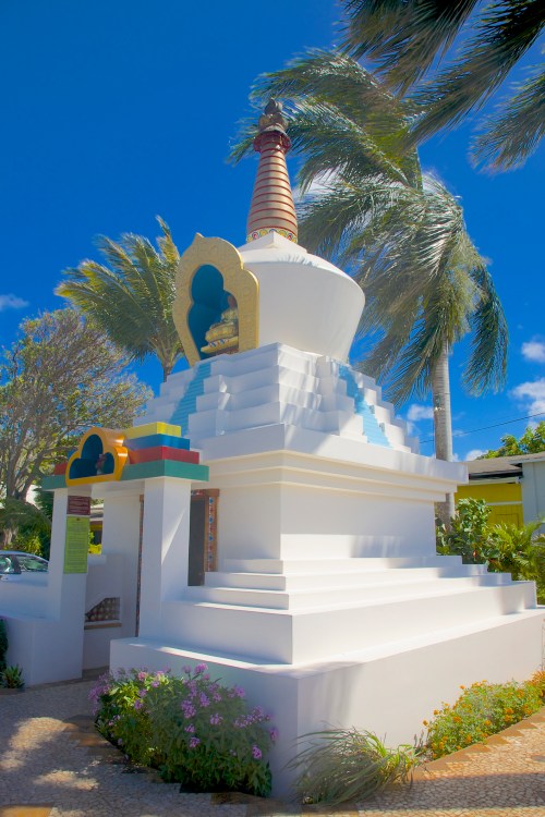 A white stupa with colorful accents, surrounded by palm trees and blue skies, set in a serene outdoor environment.