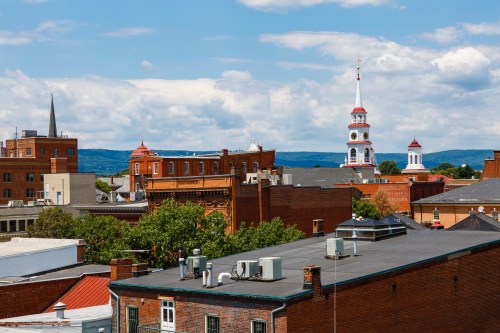 A scenic view of a town with red brick buildings, steeples, and a blue sky with fluffy clouds in the background.