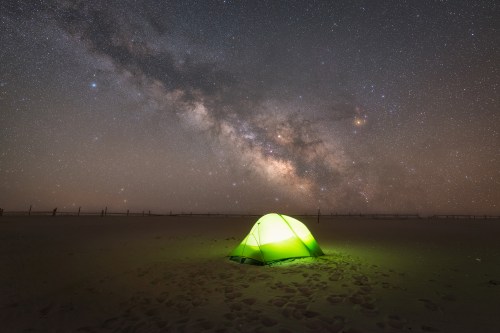 A glowing green tent on a sandy landscape under a starry night sky with the Milky Way visible.
